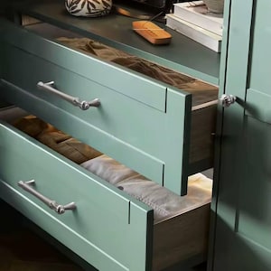 Op de afbeelding: Open lades van een mintgroene kledingkast onthullen netjes opgevouwen kleding. De lades hebben zilveren handgrepen en staan tegen een donkergroene kast. Een lamp met een patroonkap staat bovenop.