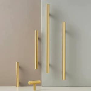 Może przedstawiać: Pięć złotych metalowych uchwytów do szafek o różnych długościach. Uchwyty są ułożone pionowo na jasnoszarym i beżowym tle.