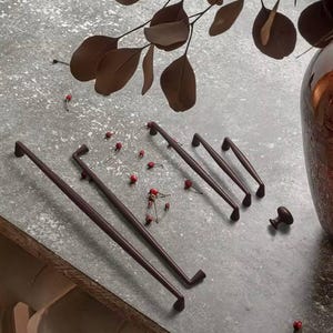 Könnte beinhalten: Eine Auswahl an dunkelbraunen Schrankbeschlägen, darunter lange Stangengriffe, kürzere Griffe und ein Knauf. Das Zubehör ist auf einer strukturierten grauen Oberfläche angeordnet, mit dekorativen roten Beeren und braunen Blättern. Der Gesamteindruck ist modern und minimalistisch.