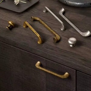 May include: A collection of cabinet hardware, including knobs and pulls, in various metallic finishes. The hardware includes a gold-toned knob, gold-toned pulls, and silver-toned pulls. The hardware is displayed on a dark wood surface.