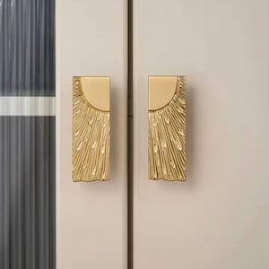 May include: Two gold-colored cabinet handles. Each handle has a rectangular base with a semi-circular top. The textured surface has a sunburst design. The handles are mounted on a light-colored cabinet door.