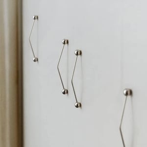 Puede incluir: Cuatro ganchos de pared de metal plateado con forma de diamante. Cada gancho tiene una pequeña bola en la parte superior e inferior. Los ganchos están montados en una pared blanca.