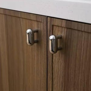Puede incluir: Dos pomos de armario plateados con forma ovalada redondeada están montados en una puerta de armario de madera.