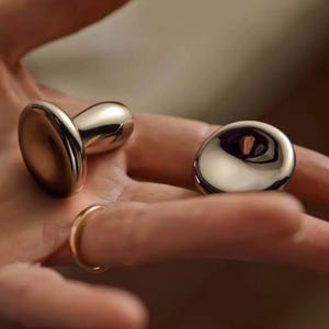 Puede incluir: Dos rodillos faciales de metal plateado. Un rodillo tiene forma de hongo con una parte superior lisa y redondeada. El otro rodillo es un disco plano con un centro ligeramente cóncavo.