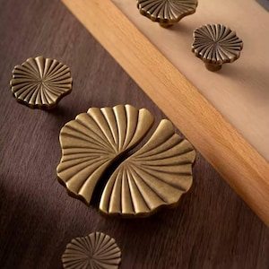 May include: Four antique brass cabinet knobs with a fan-shaped design. The knobs are arranged on a dark wood surface.