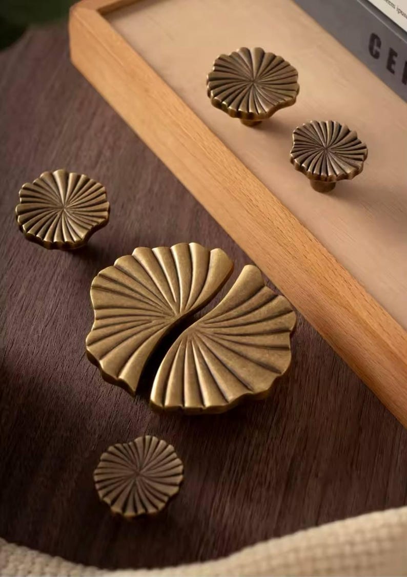 May include: Four antique brass cabinet knobs with a fan-shaped design. The knobs are arranged on a dark wood surface.