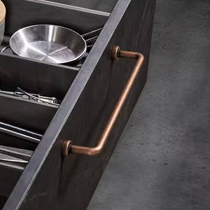 May include: Close-up of a dark gray kitchen cabinet drawer with a copper-colored handle. The drawer is open, revealing a stainless steel pan and various kitchen utensils. The handle is a straight bar with curved ends.