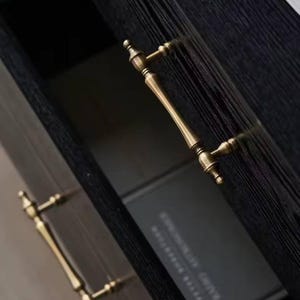May include: Close-up of a dark wooden cabinet with brass handles. The cabinet door is slightly ajar, revealing a dark interior and a book. The handles have a classic, elegant design.