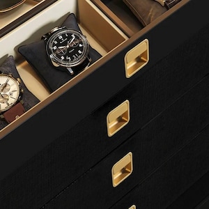 Könnte beinhalten: Schwarze Uhrenbox mit mehreren Schubladen. Jede Schublade hat einen goldenen Metall-Zuggriff. Die oberste Schublade ist geöffnet und enthält zwei Uhren.