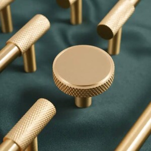 May include: A set of gold-toned cabinet knobs and pulls with a textured finish. The knobs are round with a raised edge and the pulls are rectangular with a straight edge.