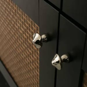 Puede incluir: Primer plano de un armario con puertas negras y paneles de ratán tejido. Las puertas del armario tienen pomos brillantes plateados de forma orgánica. El ratán es de color marrón cálido.