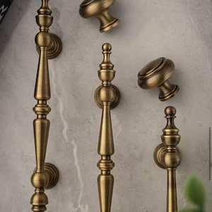 May include: Antique brass cabinet hardware set, including two long pulls and two round knobs. The pulls have a detailed, ornate design, while the knobs are simple with a rounded top. The hardware is displayed against a neutral background.
