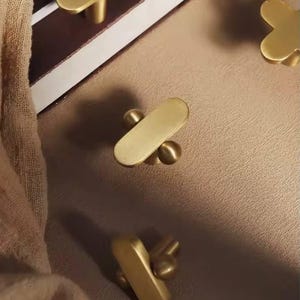 Puede incluir: Una colección de tiradores de armario de latón en varios diseños. Los tiradores incluyen una forma ovalada alargada, una forma de cruz y una forma de flor de cuatro pétalos. El hardware se muestra sobre una superficie de tono neutro.