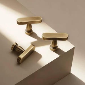 Pode incluir: Três puxadores de armário de latão antigo com design de barra em T. Os puxadores têm uma alça retangular arredondada e uma base decorativa. O hardware é exibido em uma superfície branca com um fundo neutro.