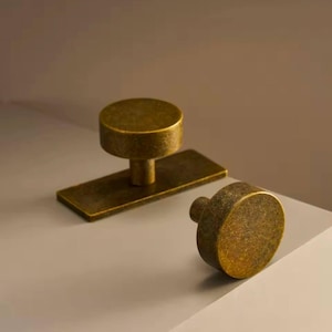 May include: Two antique brass cabinet knobs. One knob is round and has a square base. The other knob is round and has a small post.