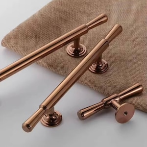 May include: Three metallic rose gold cabinet pulls of varying lengths, each featuring a cylindrical bar design with rounded ends and circular mounting bases. The hardware has a smooth, polished finish and includes a single small knob-style pull.