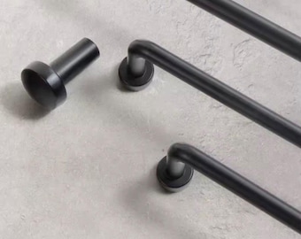 Może przedstawiać: Czarny metalowy wieszak na ręczniki o minimalistycznym designie. Wieszak posiada zakrzywione pręty i okrągłe płyty montażowe. Widoczny jest również oddzielny, cylindryczny element, prawdopodobnie pasujący.