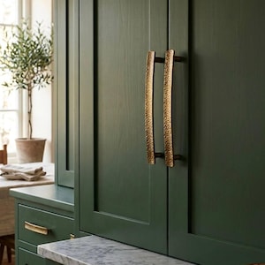 May include: Dark green kitchen cabinets with gold-toned, textured handles. The cabinets have a marble countertop and are paired with gold drawer pulls. A small potted olive tree is visible in the background, adding a touch of nature to the space.