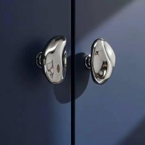 Könnte beinhalten: Zwei polierte Chrom-Schrankknöpfe mit organischer, kieselsteinartiger Form sind an einer dunkelblauen Schranktür befestigt. Die Knöpfe reflektieren die Umgebung und erzeugen einen Spiegeleffekt. Die Hardware verleiht dem Schrank eine moderne Note.