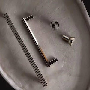 May include: A collection of contemporary metal hardware components. It features a long, rectangular bar with a mesh design, a streamlined handle, a circular knob, and a small, textured knob. The hardware is silver-toned and set on a neutral surface.