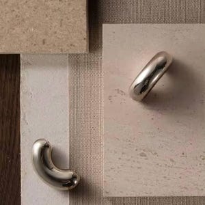Könnte beinhalten: Zwei polierte silberne Schrankgriffe, einer gebogen und einer zylindrisch, liegen auf einer neutralen Oberfläche. Die Griffe sind vor einem Hintergrund aus beige- und tanfarbenen Texturen platziert, was auf eine moderne Designästhetik hindeutet. Das Bild zeigt Hardware für die Inneneinrichtung.