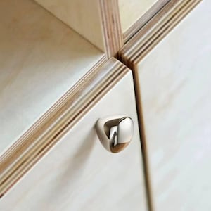 Könnte beinhalten: Nahaufnahme eines modernen Schranks mit hellem Holzmaserungsfinish. Der Schrank hat einen kleinen, abgerundeten, verchromten Griff. Die Holzmaserung ist an den Kanten des Schranks sichtbar und verleiht dem Design eine natürliche Note.