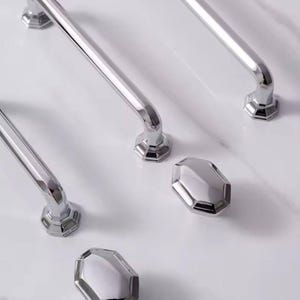 May include: A collection of polished chrome cabinet hardware, including several bar-style handles and faceted knobs. The handles have a curved design with decorative hexagonal bases. The knobs are also hexagonal and reflect light.