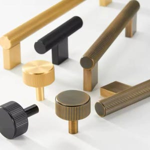 Può includere: Assortimento di pomelli e maniglie per armadi in finiture dorate e nere. I pomelli hanno un design rigato e le maniglie hanno una forma cilindrica con una texture rigata.