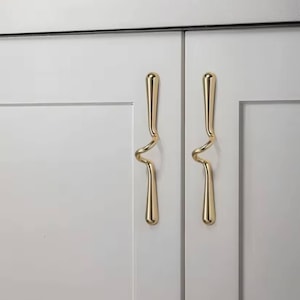 Könnte beinhalten: Nahaufnahme von zwei weißen Schranktüren mit goldfarbenen, abstrakt geformten Griffen. Die Griffe haben ein einzigartiges, geschwungenes Design, das der Schrankwand einen Hauch von moderner Eleganz verleiht. Die Griffe sind aus glänzendem Metall.