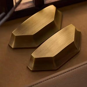 Op de afbeelding: Twee messingkleurige, geometrische kastgrepen. Elke greep heeft een facetontwerp met een rechthoekige basis en een schuine bovenkant. De grepen worden gepresenteerd op een bruin oppervlak, wat wijst op een meubel- of hardwarethema.
