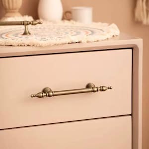 May include: Close-up of a two-drawer cabinet with antique brass hardware. The cabinet is painted a soft pink color. The drawer pulls are horizontal bars with decorative end pieces. A decorative mat and vase are on top.