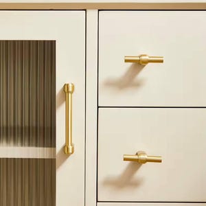 May include: A collection of gold-toned cabinet hardware, including a long bar handle and several knob-style pulls. The hardware is displayed on a cream-colored cabinet with a ribbed glass door panel. The hardware is designed for furniture.