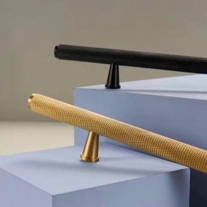 May include: Two cabinet pulls, one black and one gold, with a textured finish. Both pulls have a cylindrical shape with a tapered base.