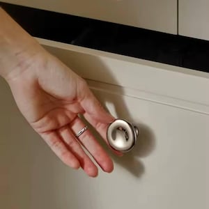 Puede incluir: Un pomo de armario de níquel cepillado con un diseño redondeado en forma de copa. El pomo está fijado a una puerta de armario de color claro. La imagen muestra un primer plano del pomo y parte del armario.