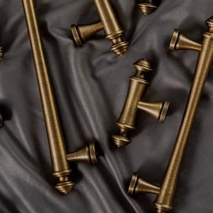 May include: A set of antique gold cabinet hardware with a variety of styles, including long pulls and smaller knobs. The hardware is displayed on a dark gray fabric background.