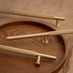 Peut inclure: Gros plan de plusieurs poignées de meubles dorées avec une texture côtelée sur la poignée. Les poignées sont montées sur un plateau en bois. Le matériel a un design moderne et minimaliste, adapté aux armoires de cuisine ou de salle de bain.