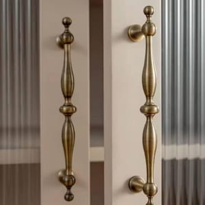 以下が含まれることがあります： 丸みを帯びた装飾的なデザインのアンティークブラス製のキャビネットドアプルが2つ。プルは白いキャビネットドアに取り付けられています。