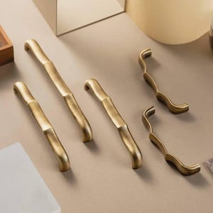 Op de afbeelding: Een verzameling messingkleurige meubelbeslag, waaronder vier handgrepen in bamboestijl en twee gebogen grepen. Het beslag is geplaatst op een neutrale ondergrond, waarbij het ontwerp en de afwerking worden benadrukt. De handgrepen hebben een textuur.