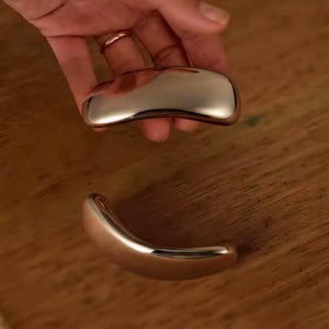 Pode incluir: Dois objetos metálicos polidos, de cor prateada, com formato oblongo e curvo. Um é segurado por uma mão com um anel dourado, e o outro repousa sobre uma superfície de madeira. Os objetos têm uma superfície lisa e reflexiva.