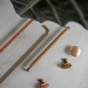 Könnte beinhalten: Eine Auswahl an Metallbeschlägen in verschiedenen Formen und Ausführungen. Das Bild zeigt gerade Stangen, gebogene Griffe und runde Knöpfe in Kupfer-, Messing- und Bronzetönen. Die Beschläge sind auf einer hellgrauen Oberfläche angeordnet.