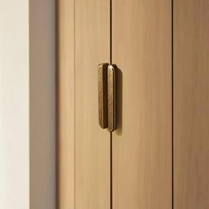 May include: A close-up shot of a wooden cabinet with a brass handle. The cabinet doors are made of light-coloured wood with vertical grain. The brass handle is rectangular with rounded edges and is mounted vertically.