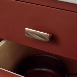 Puede incluir: Primer plano de un cajón de armario rojo con un tirador de metal cepillado. El cajón está parcialmente abierto, revelando un interior de madera y una olla oscura. Parte de un mueble de cocina o baño.