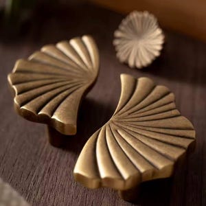 May include: Two gold-toned metal cabinet pulls shaped like a fan with a scalloped edge.
