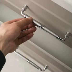 以下が含まれることがあります： テクスチャード加工されたダイヤモンド模様の中央部を備えた、研磨されたクロムキャビネットプル。プルは、各端に湾曲した直角のデザインを持っています。画像は、明るい色のキャビネットに取り付けられた2つのプルを示しています。