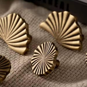 May include: Five brass cabinet knobs with a sunburst and fan-shaped design. The knobs are in various sizes and are arranged on a beige fabric surface.