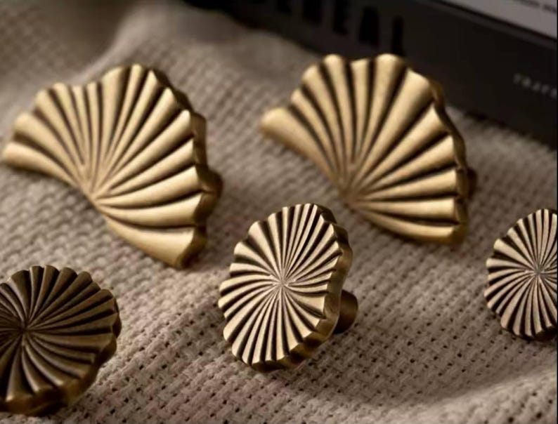 May include: Five brass cabinet knobs with a sunburst and fan-shaped design. The knobs are in various sizes and are arranged on a beige fabric surface.