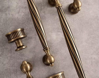 May include: An assortment of antique brass cabinet hardware, including long pulls and smaller knobs. The hardware features a ribbed design on the pulls and a decorative, rounded shape. The finish is a warm, aged brass tone.