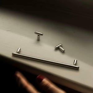 Op de afbeelding: Een set zilverkleurige kastbeslagen, bestaande uit een lange stanggreep en twee T-vormige knoppen. De hardware wordt gepresenteerd op een lichtgekleurd oppervlak, wat wijst op een moderne of minimalistische designesthetiek.