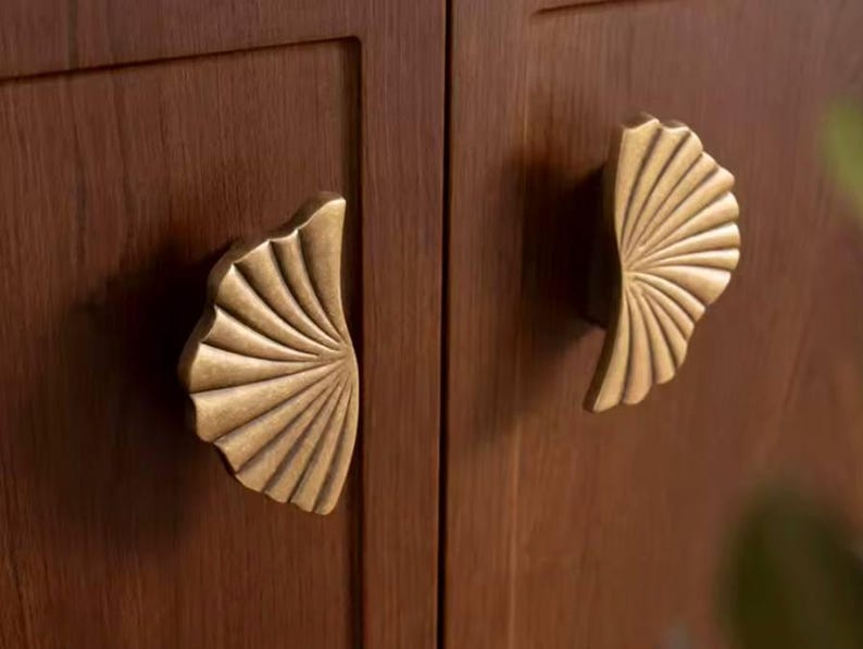 May include: Two gold-toned cabinet door pulls shaped like a fan or shell. The pulls are mounted on a wooden cabinet door.