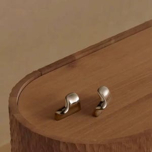 Op de afbeelding: Close-up van een houten kast met twee zilverkleurige, gebogen handgrepen. De kast heeft een getextureerd, gesneden oppervlak en een gladde, ovaalvormige bovenkant. De handgrepen zijn op het bovenoppervlak geplaatst.